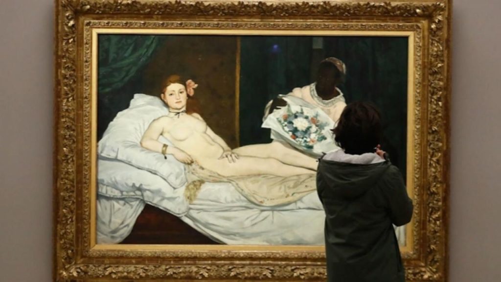 A pale young woman reclines on white sheets and pillows, completely nude except for a thin black ribbon around her neck, a gold bracelet, and a single slipper on her foot