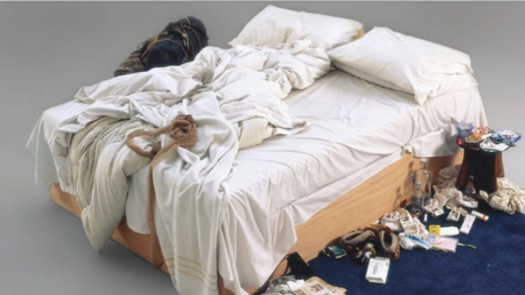 Around the bed is a constellation of personal items: ashtrays overflowing with cigarette butts (Emin was a heavy smoker), empty vodka bottles testifying to self-medication, used tissues, contraceptives, worn underwear, and slippers positioned as if just stepped out of.